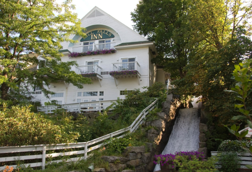 A charming white inn surrounded by greenery, featuring a small waterfall and colorful flowers in bloom.