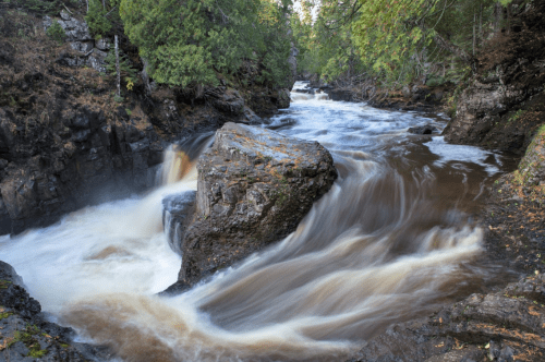 A serene river flows over rocks, surrounded by lush greenery and trees, creating a tranquil natural scene.