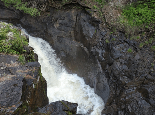 A waterfall cascading over dark rocks into a rocky gorge, surrounded by lush greenery.