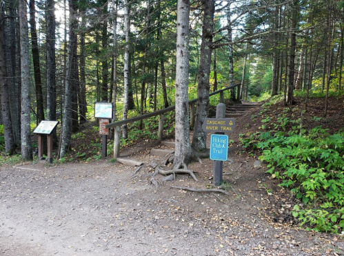 A forested trail entrance with signs for "Cascades River Trail" and "Hiking Club Trail" surrounded by trees and foliage.