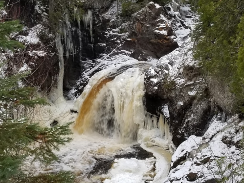 A waterfall cascades over rocky terrain, surrounded by snow and ice, with green trees in the foreground.