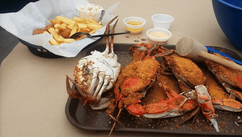 A tray of seasoned crabs with a mallet, accompanied by fries and dipping sauces in the background.