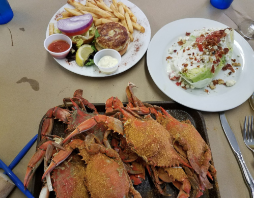A platter of seasoned crabs with fries, a burger, and a wedge salad topped with bacon and dressing on a table.