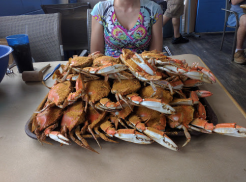 A large platter piled high with seasoned crabs, with a person in a colorful shirt sitting behind it.
