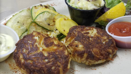Two golden crab cakes served with sautéed zucchini and squash, a side of coleslaw, and dipping sauces.