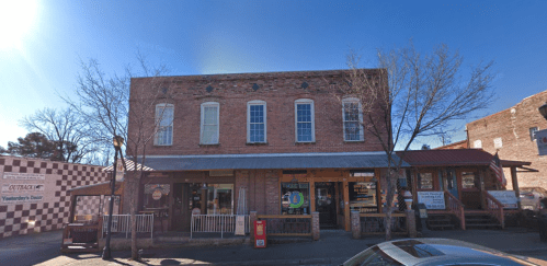 A brick building with large windows, a covered porch, and nearby shops on a sunny day.