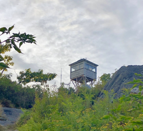 A wooden lookout tower perched on a rocky hill, surrounded by greenery under a cloudy sky.