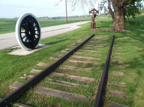 A grassy area with old railway tracks, a large wheel sculpture, and a wooden figure near a road and fields.