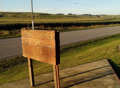 Sign for the Jesse James Historical Site, with fields and a road in the background, under a clear sky.