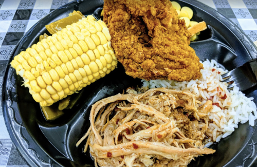A black plate with fried chicken, corn on the cob, shredded pork, and rice, set on a checkered tablecloth.