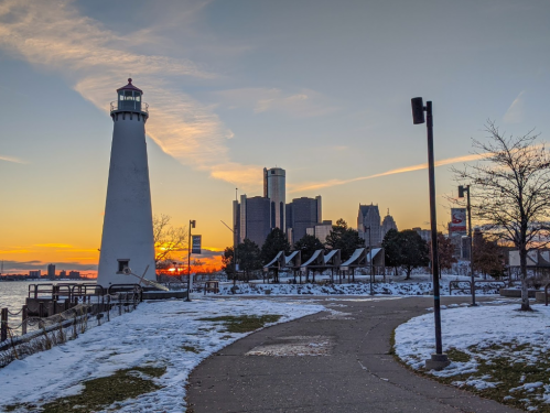 A lighthouse stands by a snowy path with a city skyline at sunset in the background.