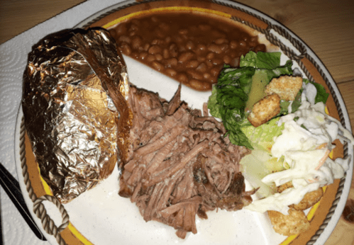 A plate featuring pulled pork, a baked potato wrapped in foil, baked beans, and a side salad with croutons.
