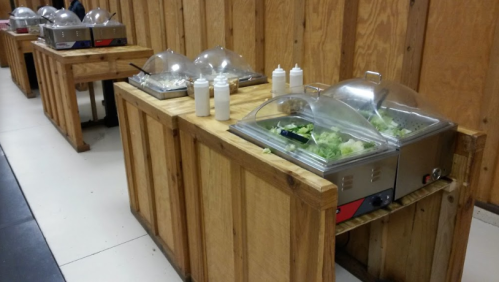 Buffet setup with wooden tables, covered food trays, and condiment bottles in a casual dining area.