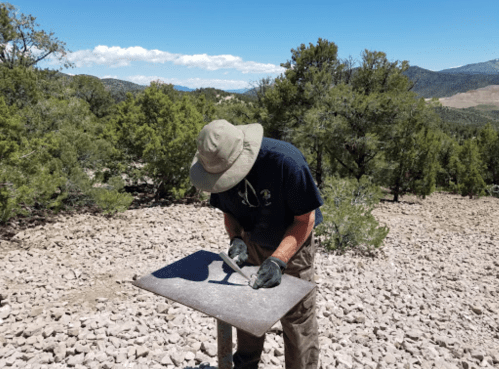 A person in a hat and gloves chisels a stone tablet in a rocky, mountainous area with trees and blue sky.