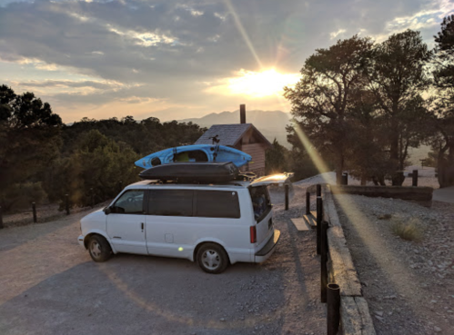 A van parked at a scenic spot with a kayak on top, surrounded by trees and a sunset in the background.