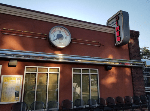 Exterior of a diner with a large clock and neon sign, featuring a menu board and outdoor seating.
