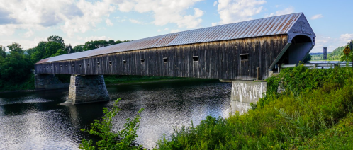 A long wooden covered bridge spans a calm river, surrounded by greenery and under a blue sky with scattered clouds.