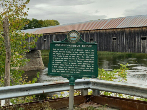 Sign for the Cornish-Windsor Bridge, the longest wooden covered bridge in the U.S., with a river and wooden structure in the background.