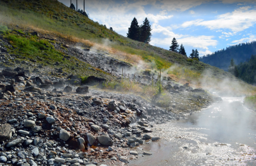 Steam rises from a rocky riverbank surrounded by lush greenery and mountains under a blue sky.