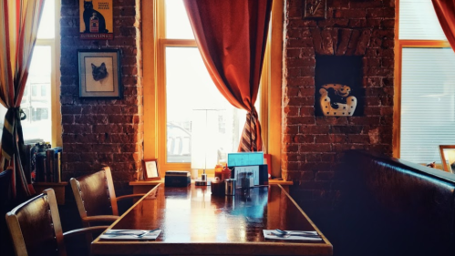 A cozy restaurant interior with a wooden table, chairs, and colorful curtains by large windows.