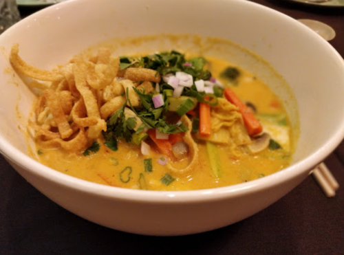A bowl of curry soup topped with vegetables, herbs, and crispy noodles.