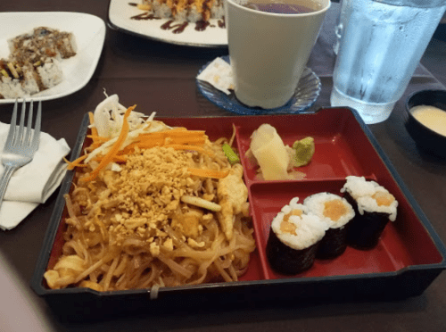A red tray with pad thai topped with peanuts, sushi rolls, pickled ginger, and wasabi, alongside a cup of tea and water.