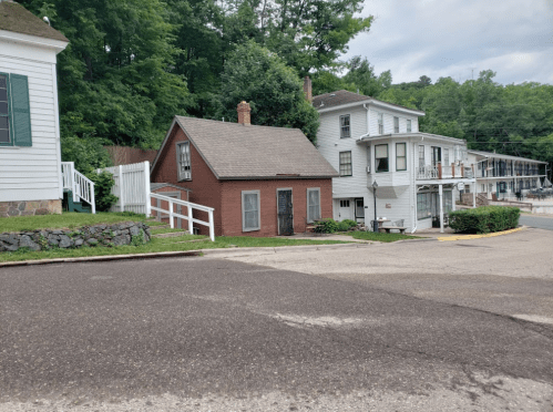 A small red house with a sloped roof, surrounded by greenery, next to a larger white building on a quiet street.