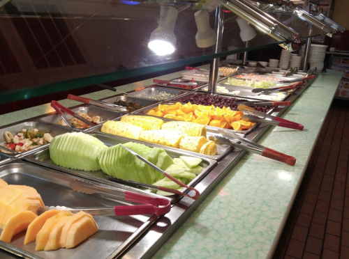 A buffet display featuring a variety of fresh fruits and vegetables in metal trays with tongs for serving.