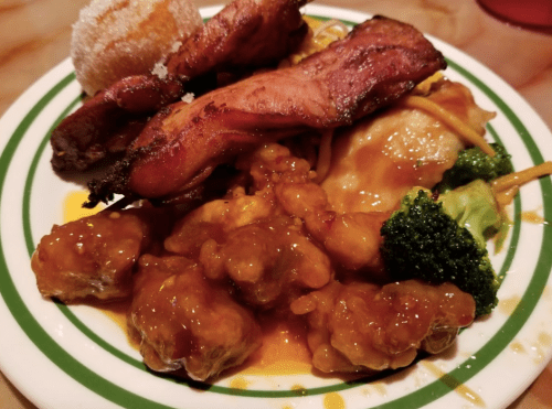 A plate of food featuring grilled meat, sweet and sour chicken, broccoli, noodles, and a dessert.