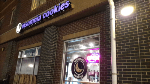 Exterior of Insomnia Cookies shop at night, featuring a lit sign and window displaying cookie offerings.