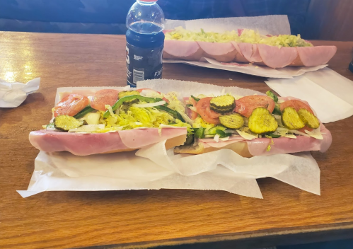 Two large sandwiches filled with ham, lettuce, tomatoes, pickles, and green peppers, served on paper with a soda in the background.
