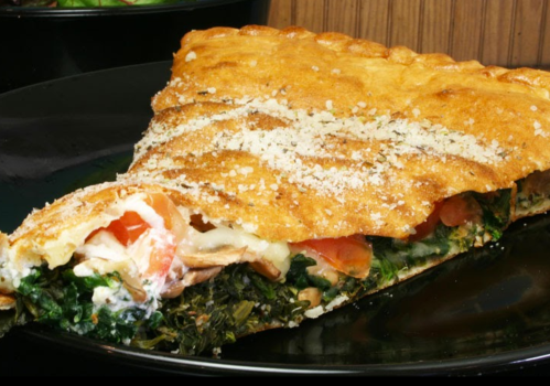 A slice of golden-brown savory pie filled with greens, tomatoes, and cheese, served on a black plate.