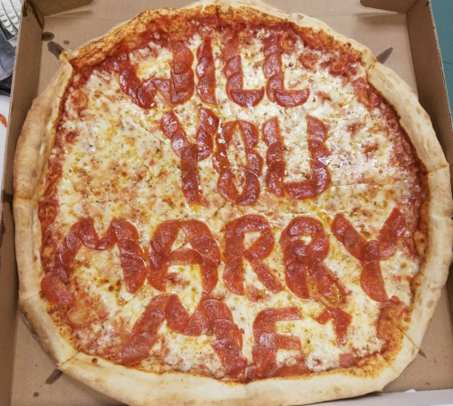 A large pizza with melted cheese and pepperoni arranged to spell "Will you marry me?" on the surface.
