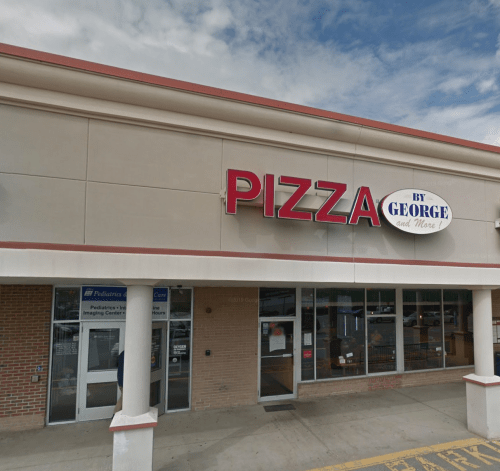 Exterior of a pizza restaurant named "Pizza by George" with a blue sky and nearby storefronts visible.