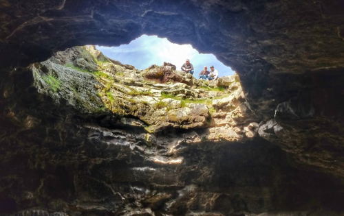 View from inside a cave looking up at two people sitting on the rocks above, with bright sky visible through the opening.