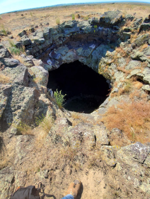 A rocky, circular opening in the ground surrounded by dry grass and stones, revealing a dark abyss below.