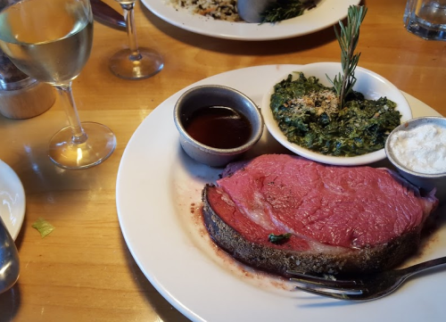 A plate featuring a slice of prime rib, sides of spinach, sauce, and a glass of white wine on a wooden table.