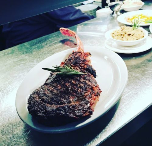 A beautifully cooked ribeye steak garnished with rosemary, served on a white plate with sides in the background.