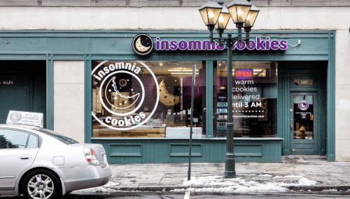 Exterior of Insomnia Cookies shop with a sign, open until 3 AM, and a car parked nearby on a snowy street.