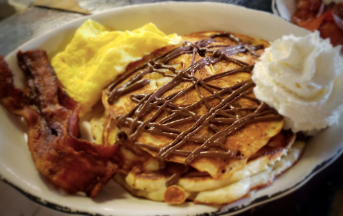 A plate of pancakes drizzled with chocolate, served with scrambled eggs, bacon, and whipped cream.