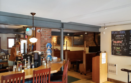 A cozy café interior with wooden tables, a counter, and a chalkboard menu. A barista is preparing drinks.