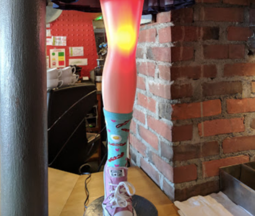 A colorful leg lamp with a glowing knee, wearing fun socks and a sneaker, stands against a brick wall in a café.