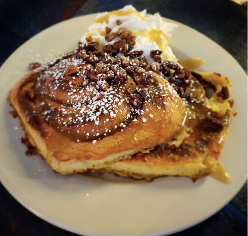 A plate of French toast topped with pecans, powdered sugar, and a drizzle of syrup, served with whipped cream.