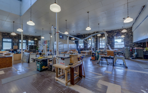 A spacious workshop with various woodworking tools, large windows, and overhead lights illuminating the area.