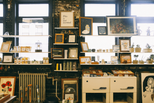 A cozy shop interior with shelves displaying art, plants, and various decorative items against a brick wall.