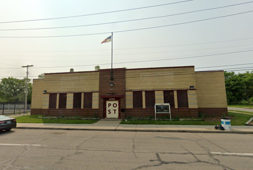 A brick building with a flag, labeled "POST," located on a street with power lines overhead.