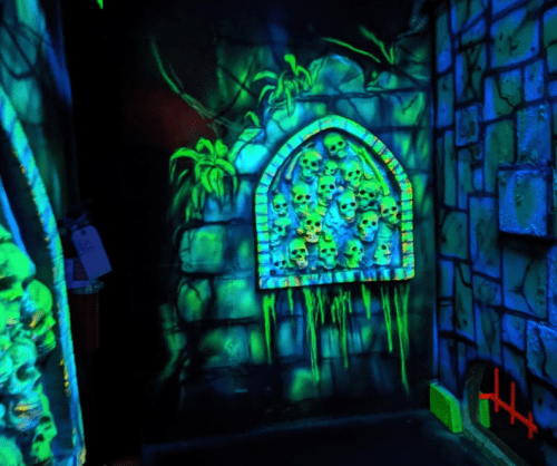 A dark, glowing room with a stone wall featuring a niche filled with skulls and green dripping slime.