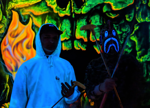 Two individuals stand in front of a glowing skull mural, one wearing a white hoodie and the other in a mask, holding golf clubs.