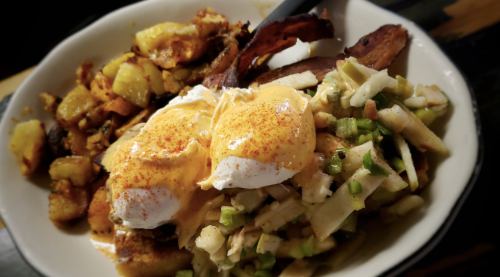 A bowl of poached eggs topped with hollandaise sauce, served with potatoes and bacon on the side.