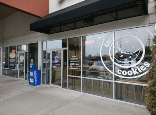 Exterior of Insomnia Cookies shop with large windows, open sign, and a delivery notice. Bright and inviting atmosphere.